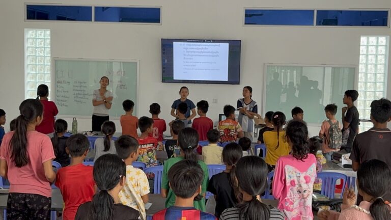 Older students standing at the front of a classroom, leading a lesson for rows of younger children